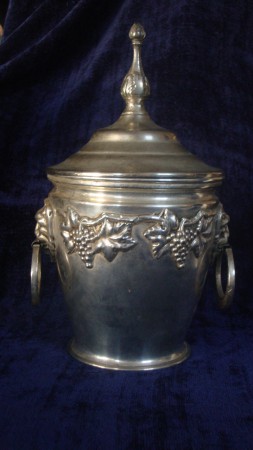 Balde de gelo em casquinha com sinais de banho de prata. Apresenta decoração com cachos de uva e cabeças de leão a suspender as argolas.